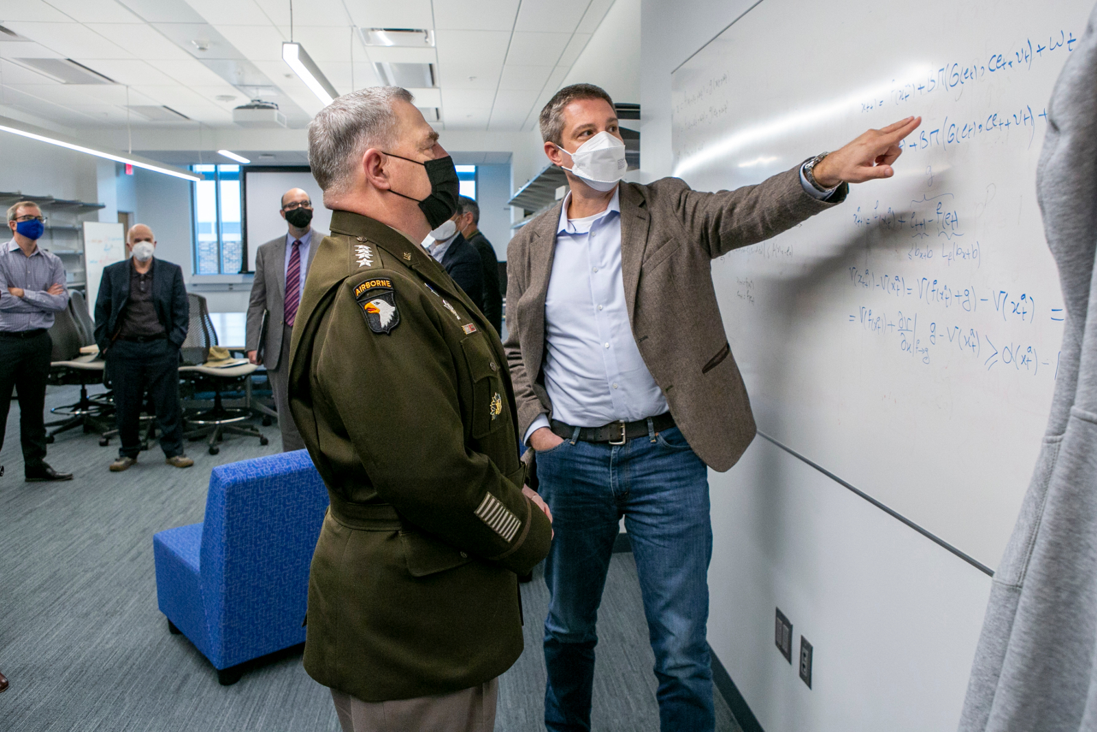 A general stands with a man at a white board who is pointing to equations written on it