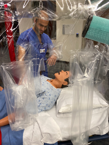 A medical caregiver stands next to a dummy on a bed under a plastic tent