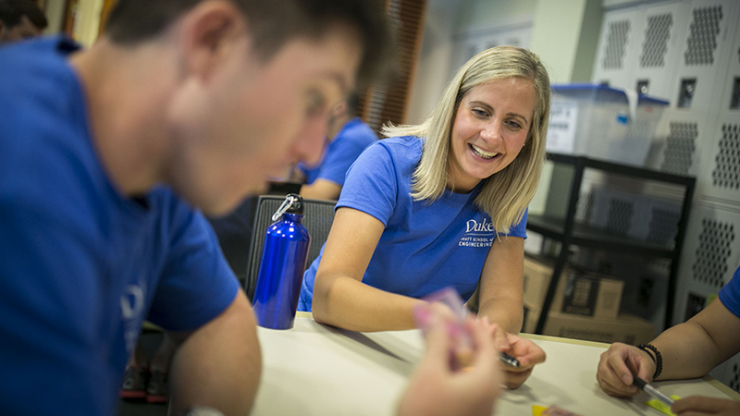 A smiling blond woman in a Duke blue t-shirt works at a table with a younger man