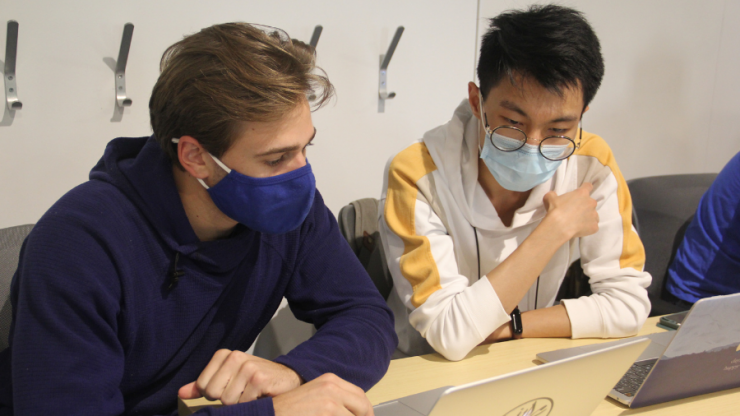 Two students in masks look at laptops in a classroom