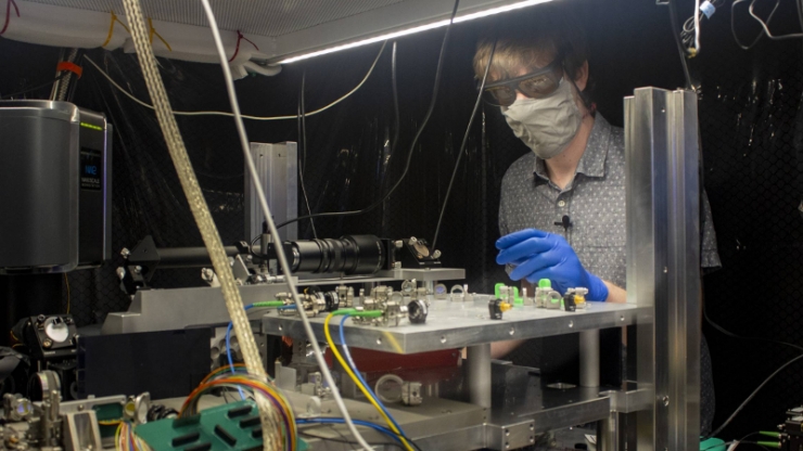 A man in a mask and gloves works at a bench with a lot of electrical and optical equipment