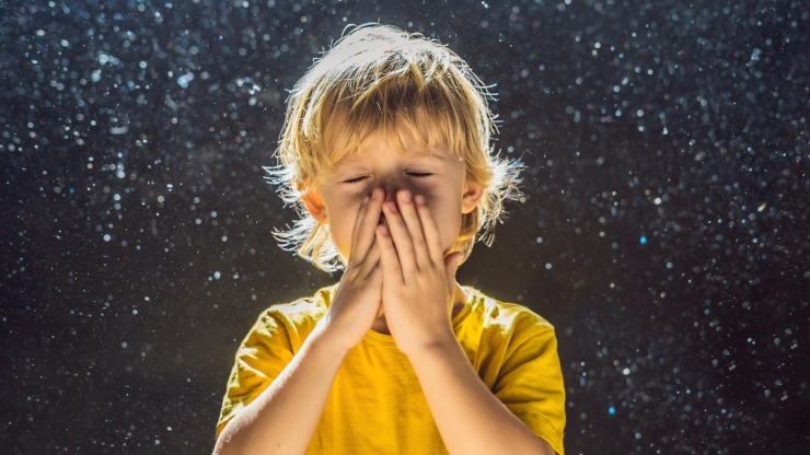A blond boy in a yellow t-shirt sneezing in a cloud of dust