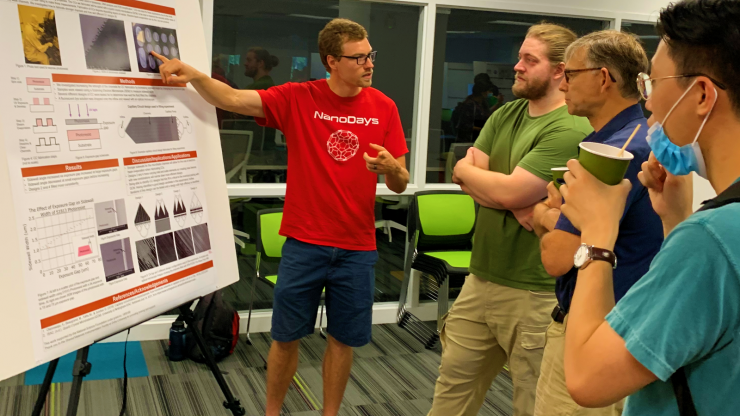 man in red shirt pointing at research poster on easel and explaining it to three other men