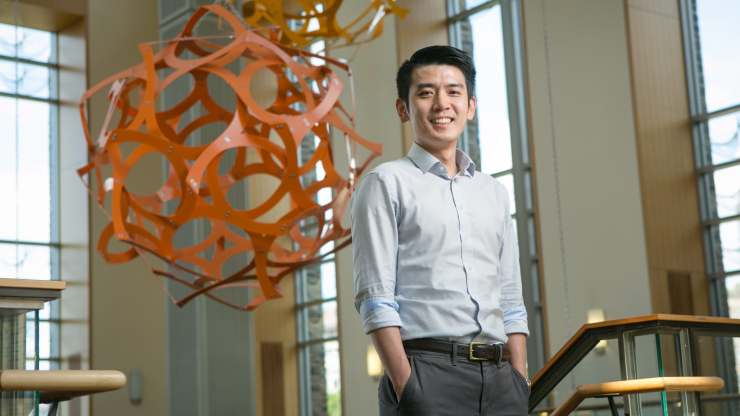 Po-Chun Hsu standing with hands in pockets at top of stairs with hanging orange sculpture in background