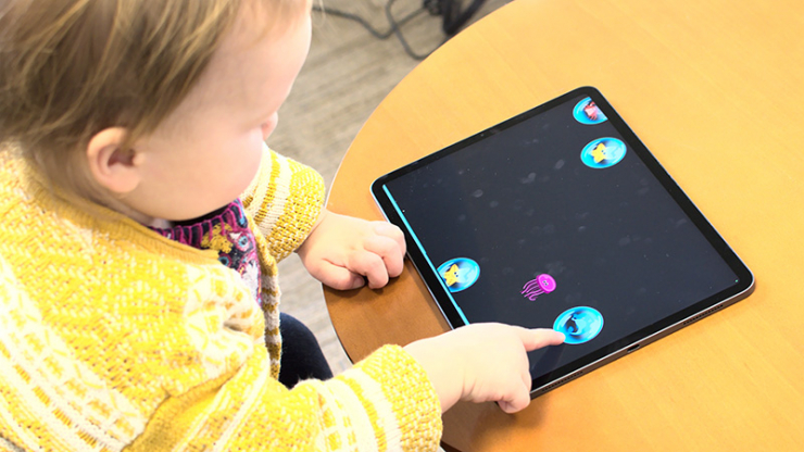 A young child at a table in a yellow shirt pointing a finger at a bubble on a tablet screen