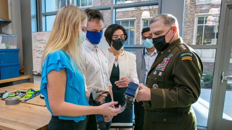A man in a general jacket speaks with a team of students holding a belt-like object