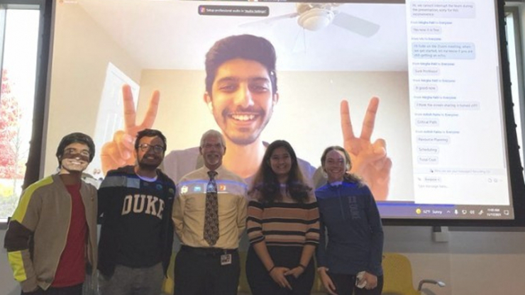 A teacher and students standing in front of a projector screen featuring a student on zoom