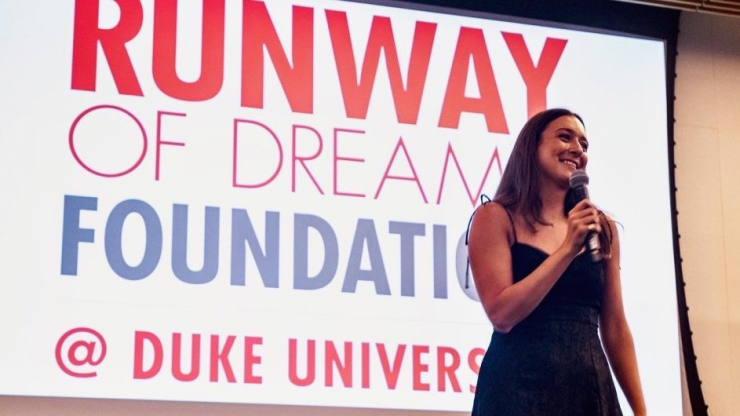 woman with long dark hair holding microphone on stage with Runway of Dream Foundation @ Duke University sign as backdrop