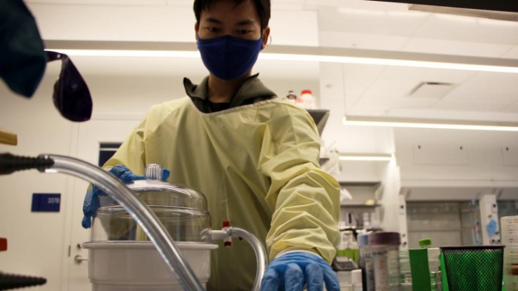 A gloved, gowned and masked man reaches toward a centrifuge and samples in vials
