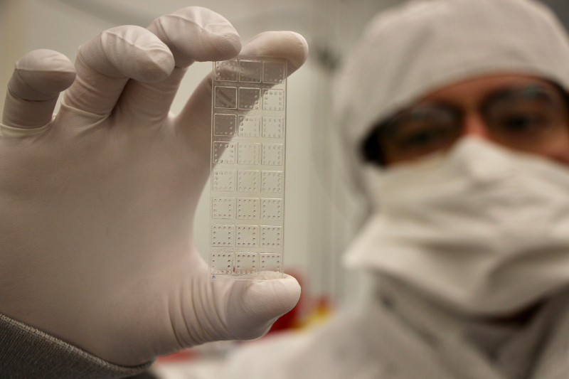 A scientist in a clean room coat holds a small rectangular plastic slide with lots of squares