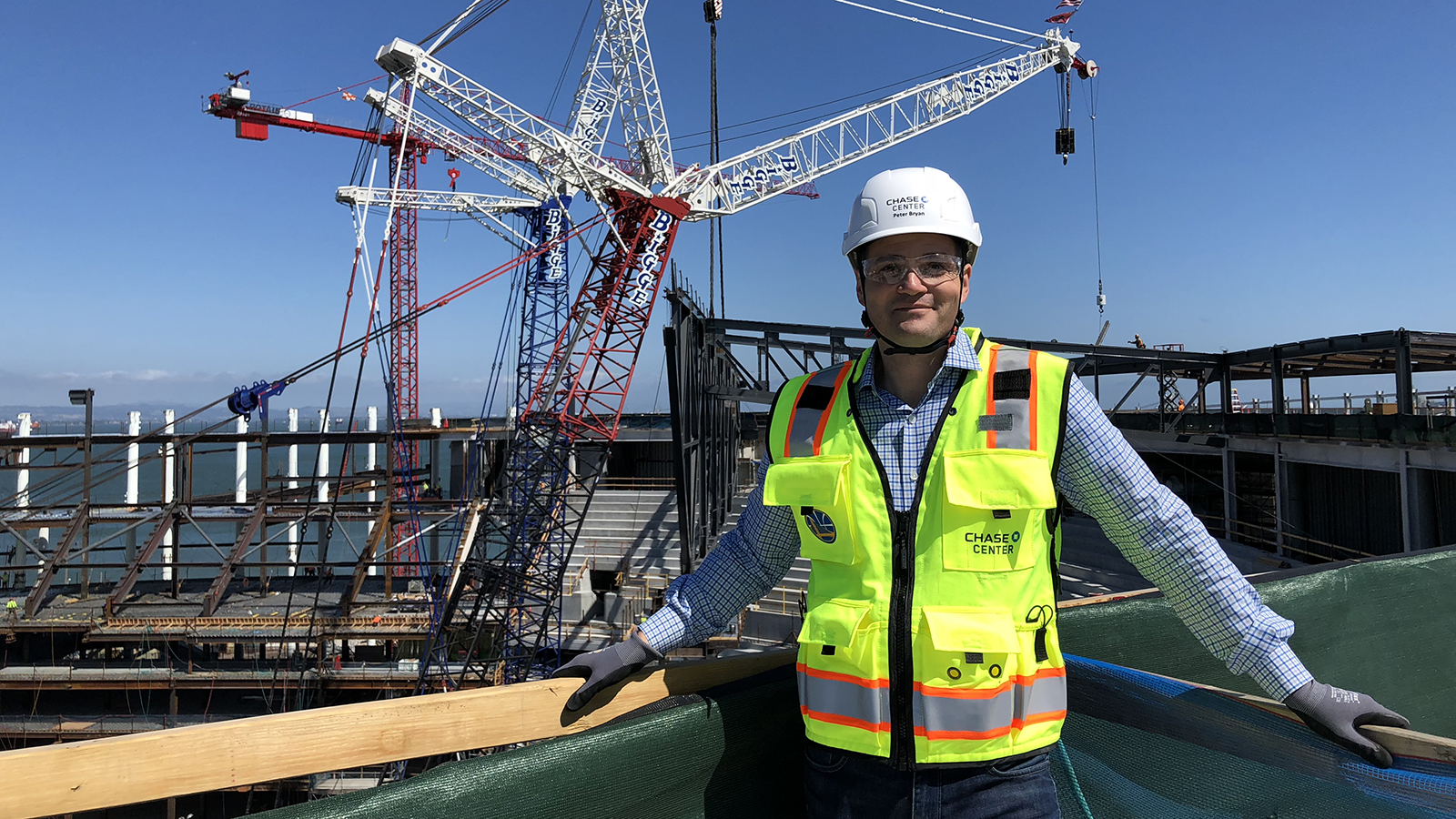 A man stands with a white hard hat and a neon green safety vest in front of a large multi-armed crane over water