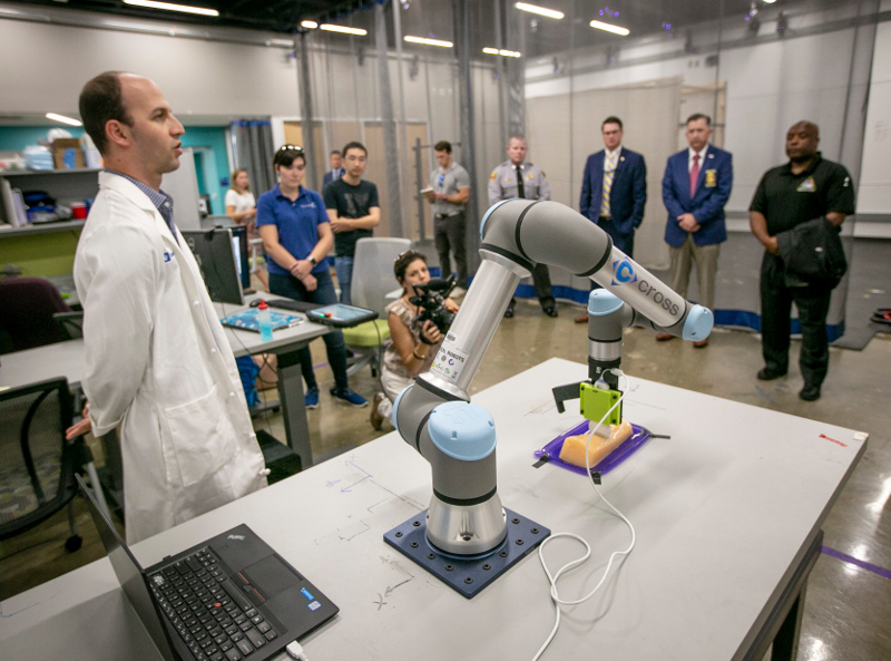 A man in a white lab coat standing by a robotic arm in front of a group of people
