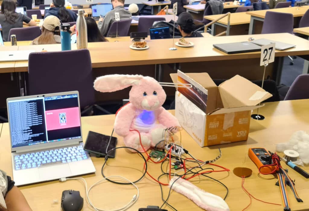 a pink bunny sits on a desk in a crowded lecture hall with a computer and wires everywhere