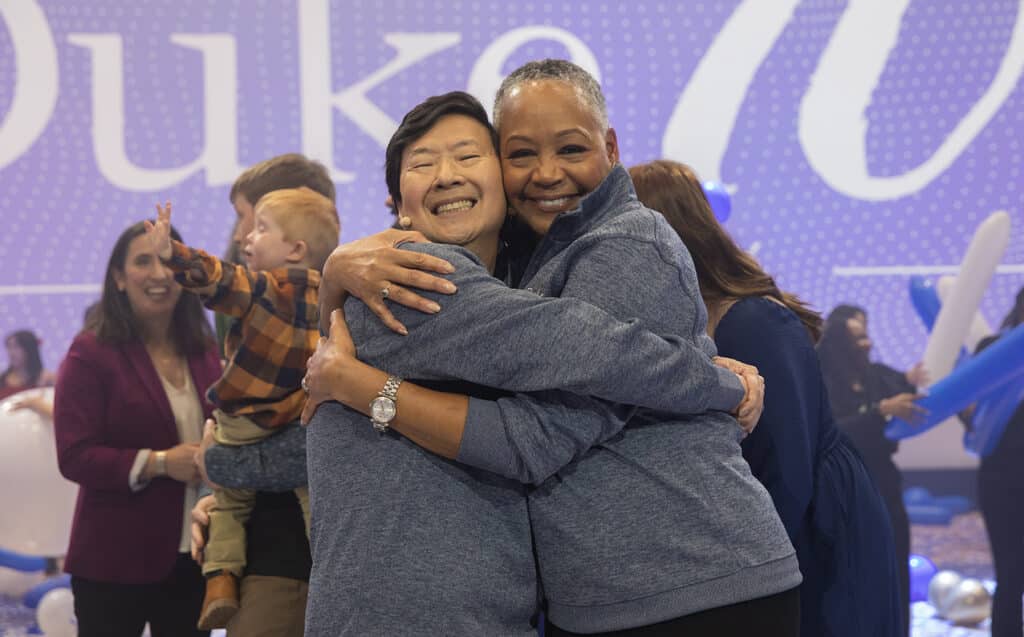 A man and a woman hug with lots of people and balloons in the background.