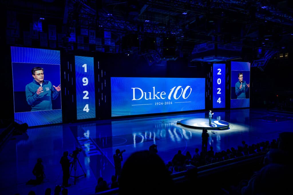 A man stands with a microphone in dim blue lights on a basketball court with huge screens around him.