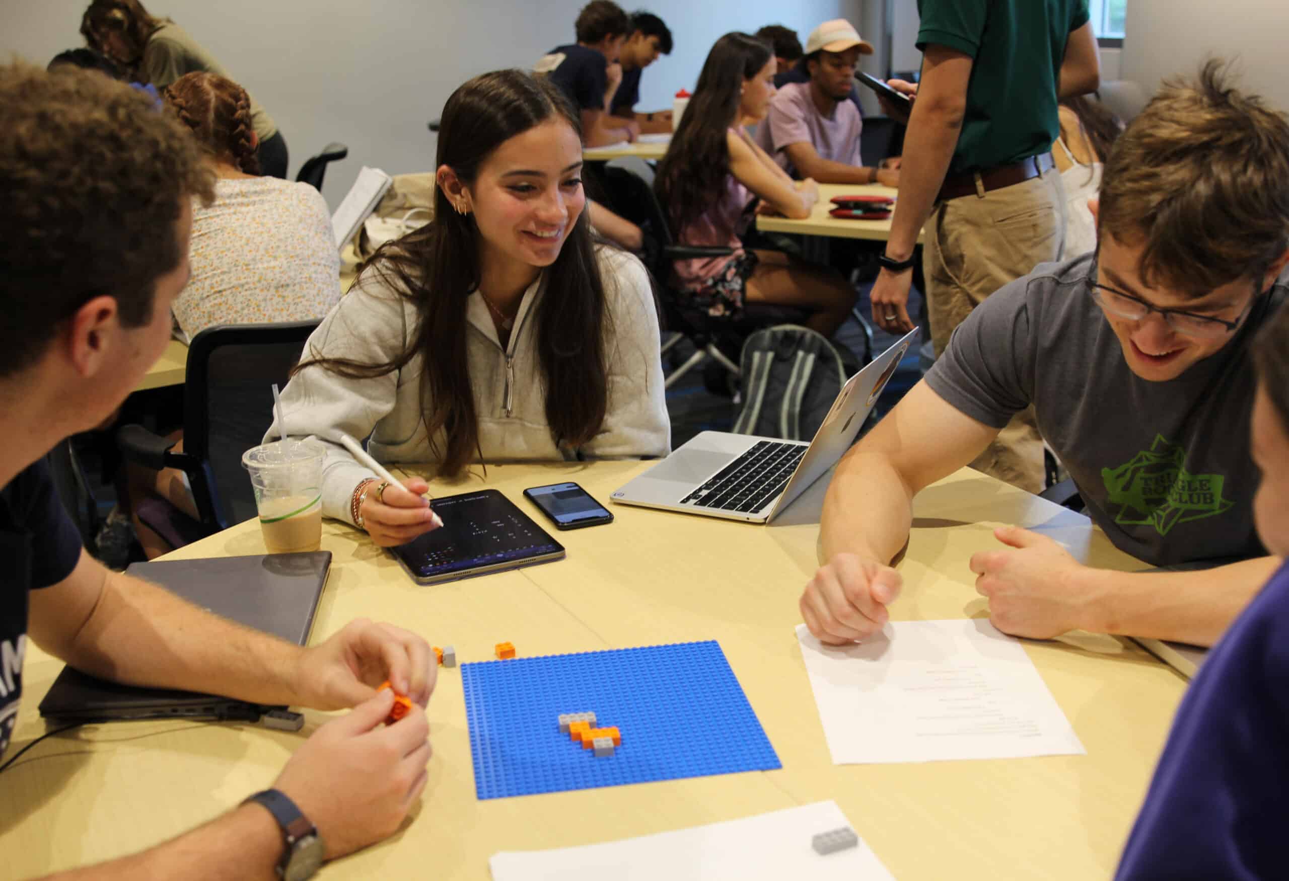 A group of three students sit around a piece of paper and board with Lego pieces.