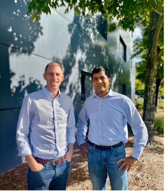 Both in light blue dress shirts and jeans, Reifschneider and Singh stand outside a building under a tree in sunlight, looking at the camera.