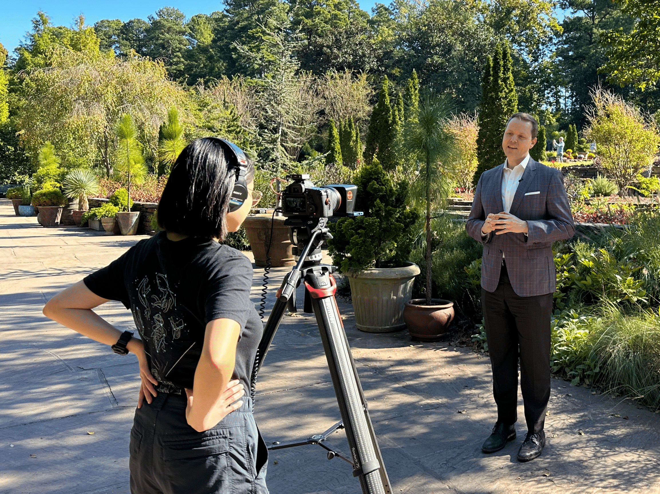 Maddie standing behind a camera, filming Dean Jerry who is standing in front of the camera. The Duke gardens full of green foliage is in the background.