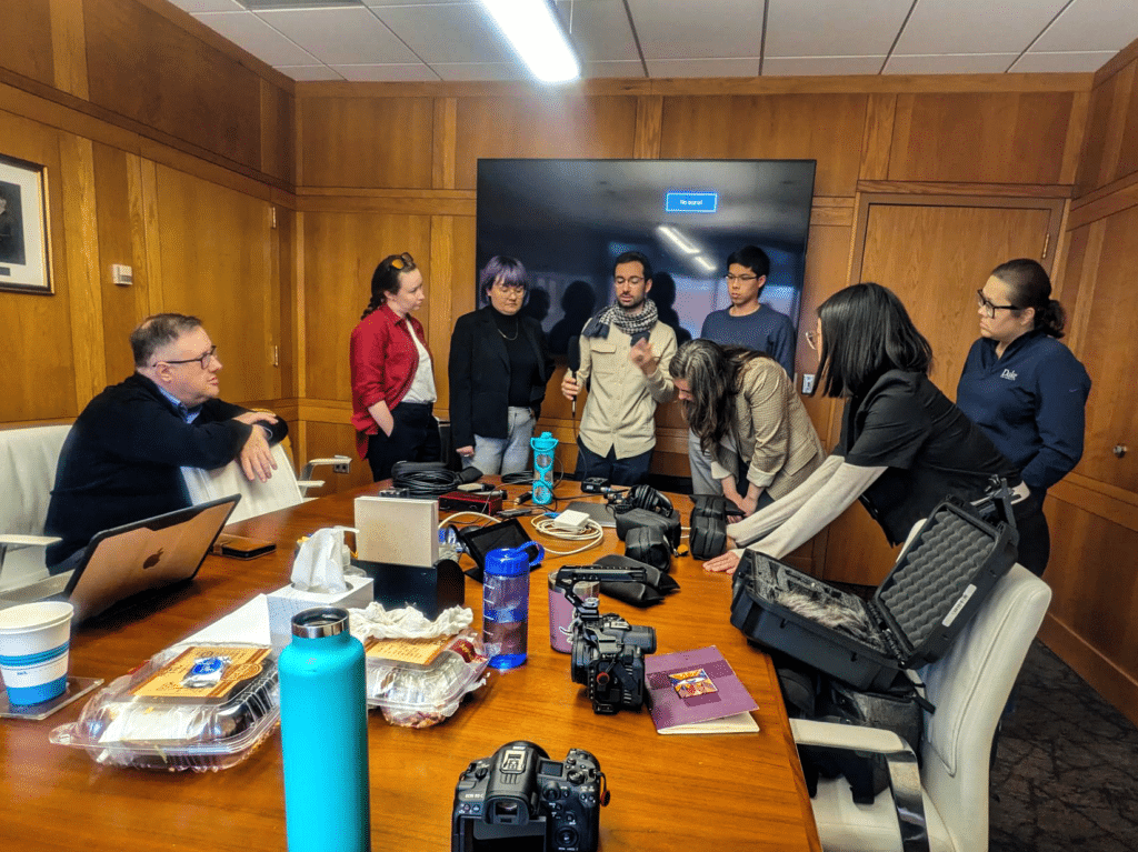 A group of 8 people standing around a meeting room table that's covered in camera and audio equipment. They are all listening the man in the center with glasses, who is teaching them about audio.