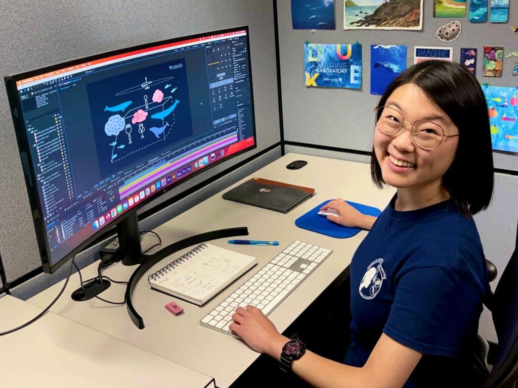 Maddie smiling at the camera while working at a monitor at her desk. The monitor shows illustrations of whales and other sea animals. Behind Maddie is a wall of decorative posters and prints, one of which contains the words "Duke Marine Laboratory."