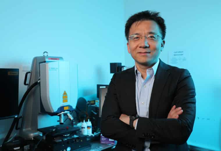Man in glasses standing with arms crossed in front of a microscope in a lab.