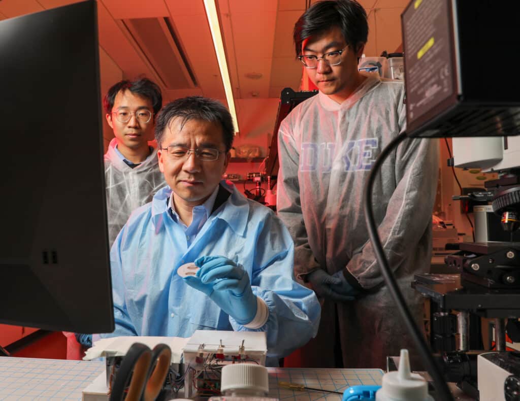 Two men stand behind a man seated at a lab bench. All three men are looking at a white circular device held in the seated man's gloved hand.