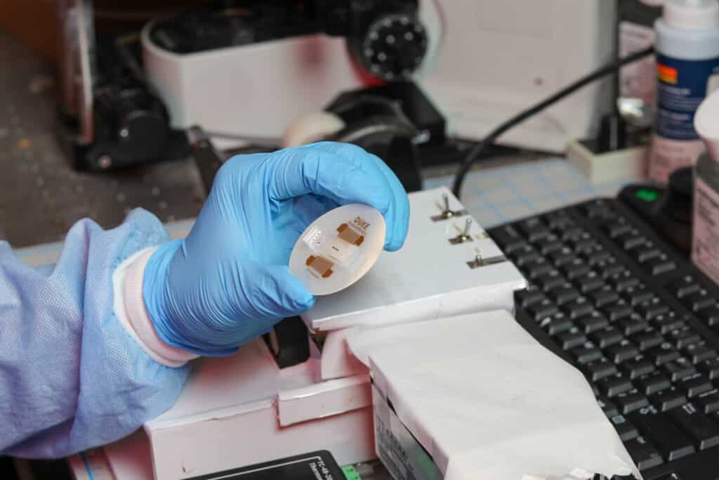 A blue gloved hand holds a white circular device around the size of a petri dish with rectangular electronics on it. On the device are printed the words "Duke Acoustofluidics" and "CTC Separation."