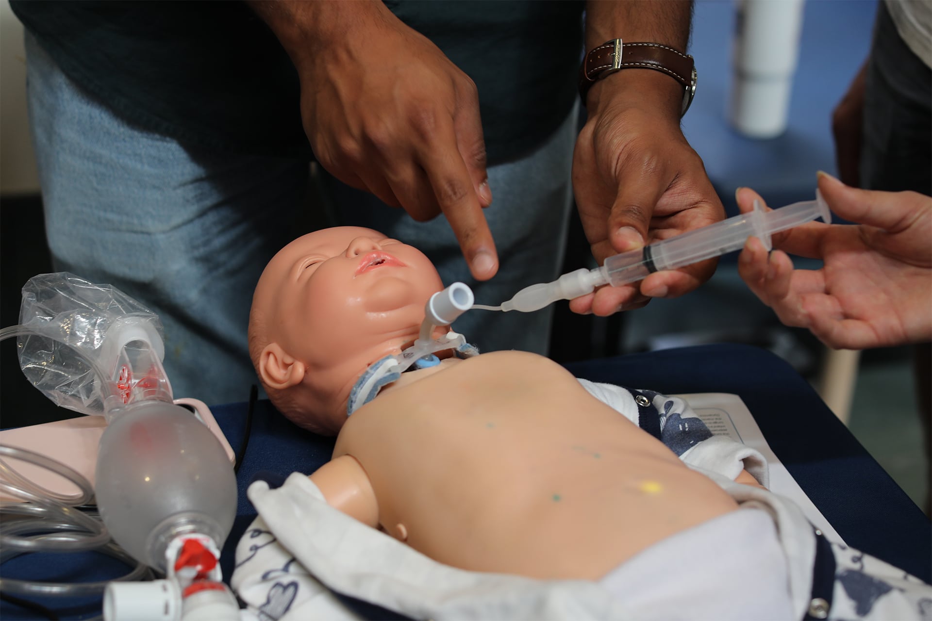 A baby manikin with a trach tube in its neck lying face up on a table. Two people are connecting a plastic syringe to the open end of the manikin's trach tube. Other related hospital equipment can also be seen on the table.