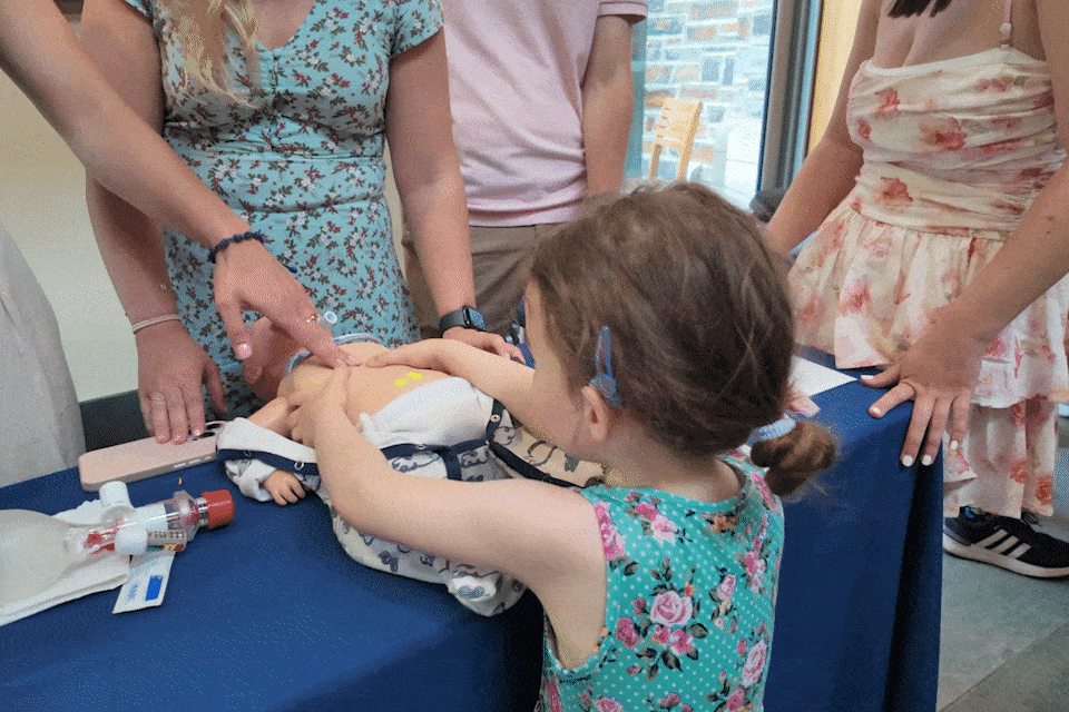 A small child practices giving CPR compressions to a manikin baby with help from a college student.