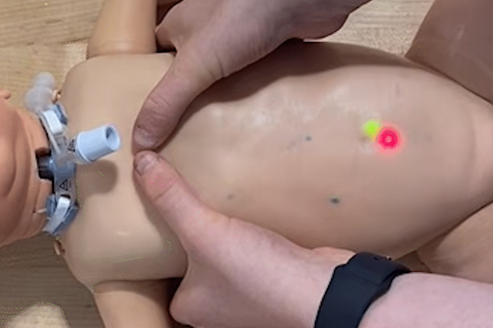 Closeup of person doing CPR on a baby manikin. A red light and then a yellow light appear on the manikin's belly.