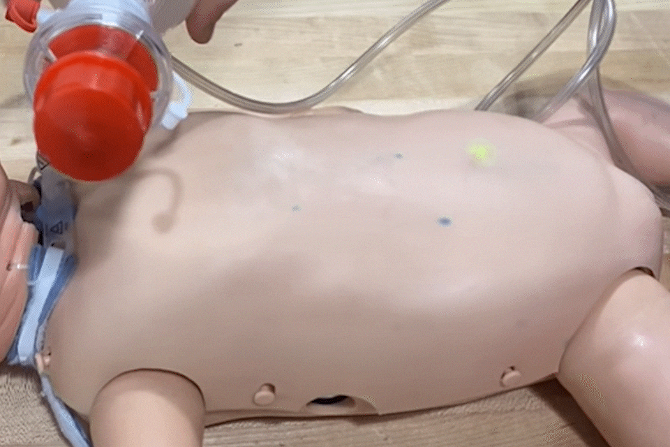 A person squeezes an air bottle connected to the neck of a baby manikin. The manikin's chest rises and falls as the air bottle is squeezed and released.