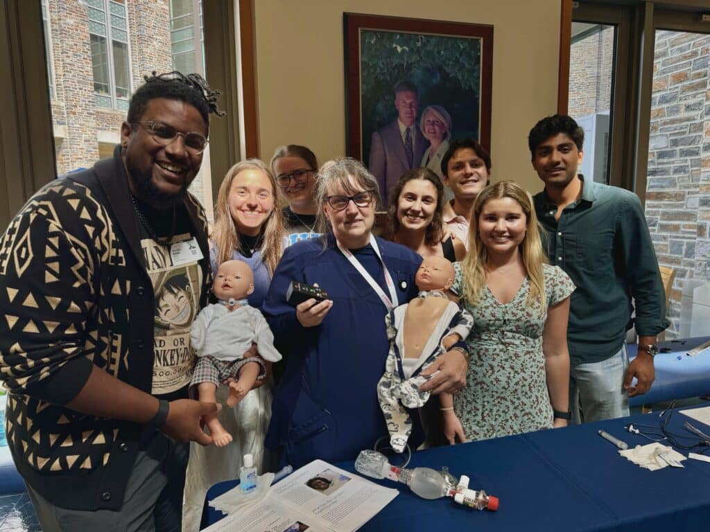 A group of eight people smile for a group photo. Two of the people hold baby manikins with trach tubes in their necks.