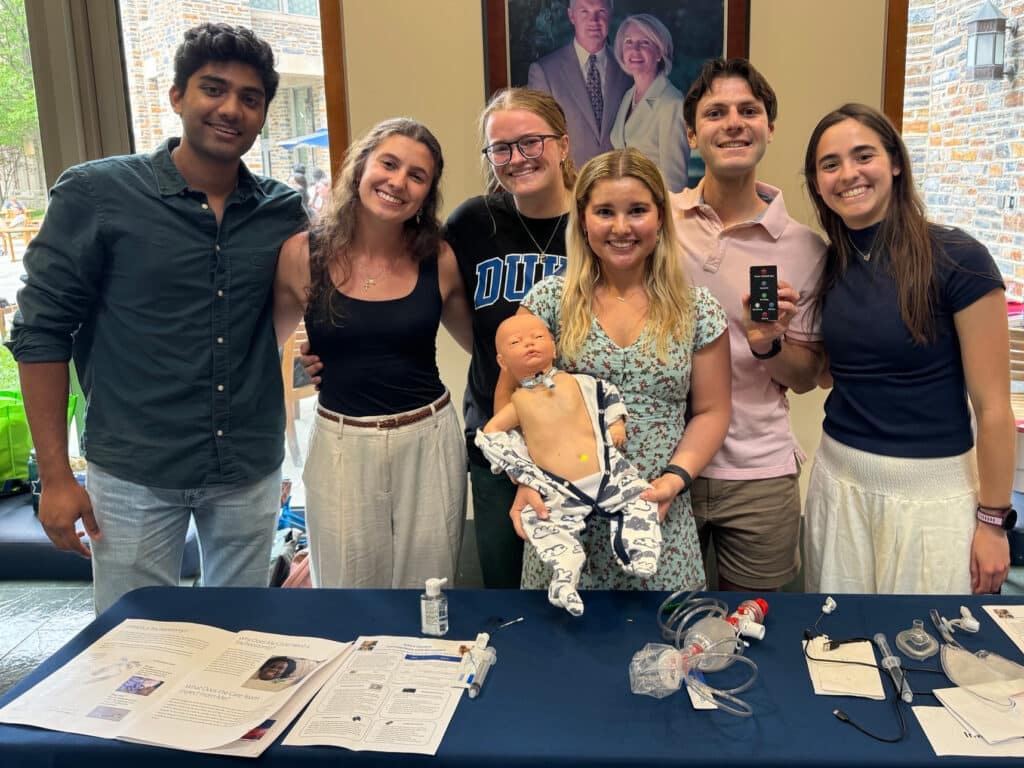 Six college students smiling for a group photo. One of the center students is holding a baby manikin with a trach tube, and another is holding up a remote. control.