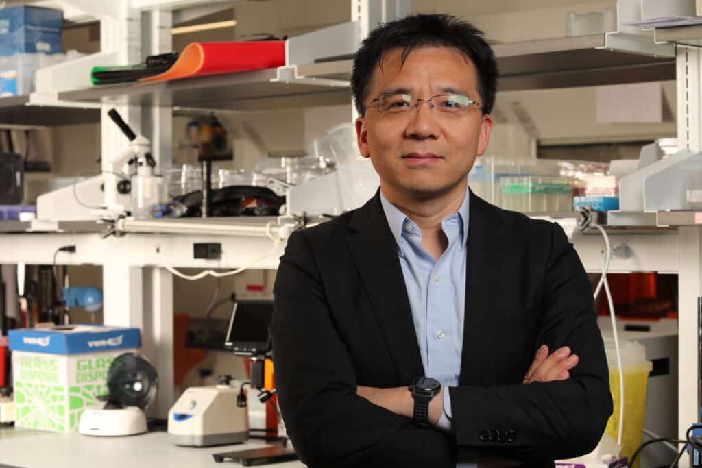 Man in glasses standing with arms crossed in front of a lab bench,