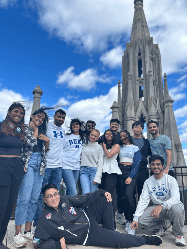 A group of students poses together in front of a pointed spire belonging to Duke Chapel under a bright blue sky with scattered clouds. One student is reclining playfully in front while others stand closely behind, smiling.