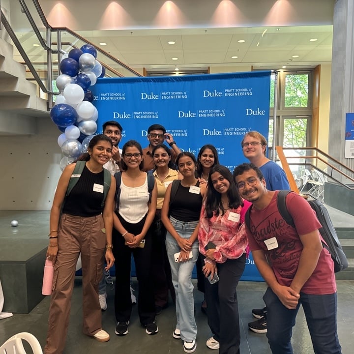 A group of students stands indoors in front of a Duke Pratt School of Engineering backdrop decorated with blue and white balloons. They are smiling and posing for a group photo, some holding backpacks and water bottles.