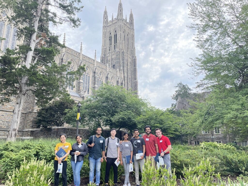 A group of students stands in front of Duke Chapel, with its tall Gothic-style tower rising behind them. The students are smiling, holding folders and bags, surrounded by greenery and trees on campus.