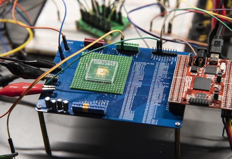 Close-up of a laboratory electronics test setup with a circuit board, prototype microchip, colorful wires, and connected testing equipment.