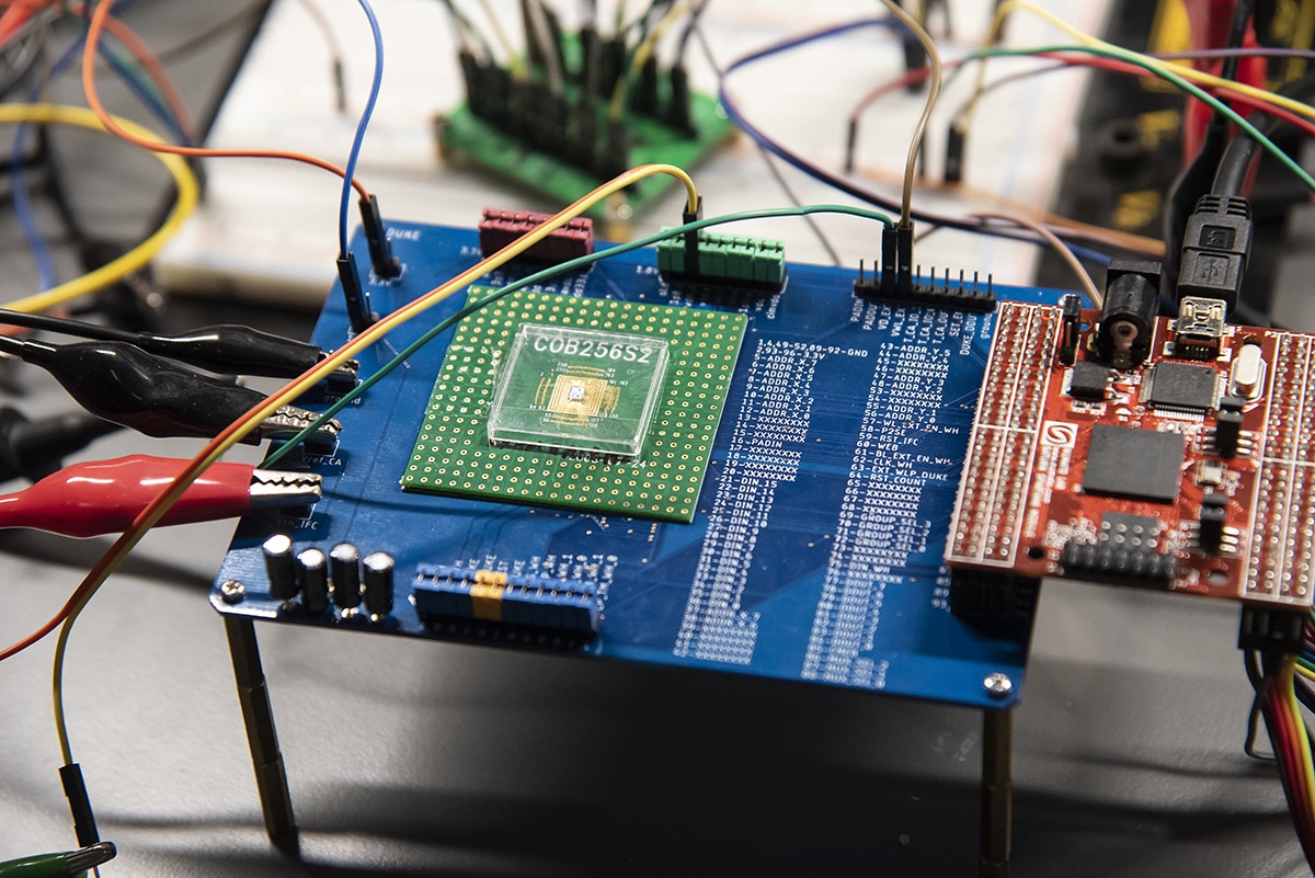 Close-up of a laboratory electronics test setup with a circuit board, prototype microchip, colorful wires, and connected testing equipment.